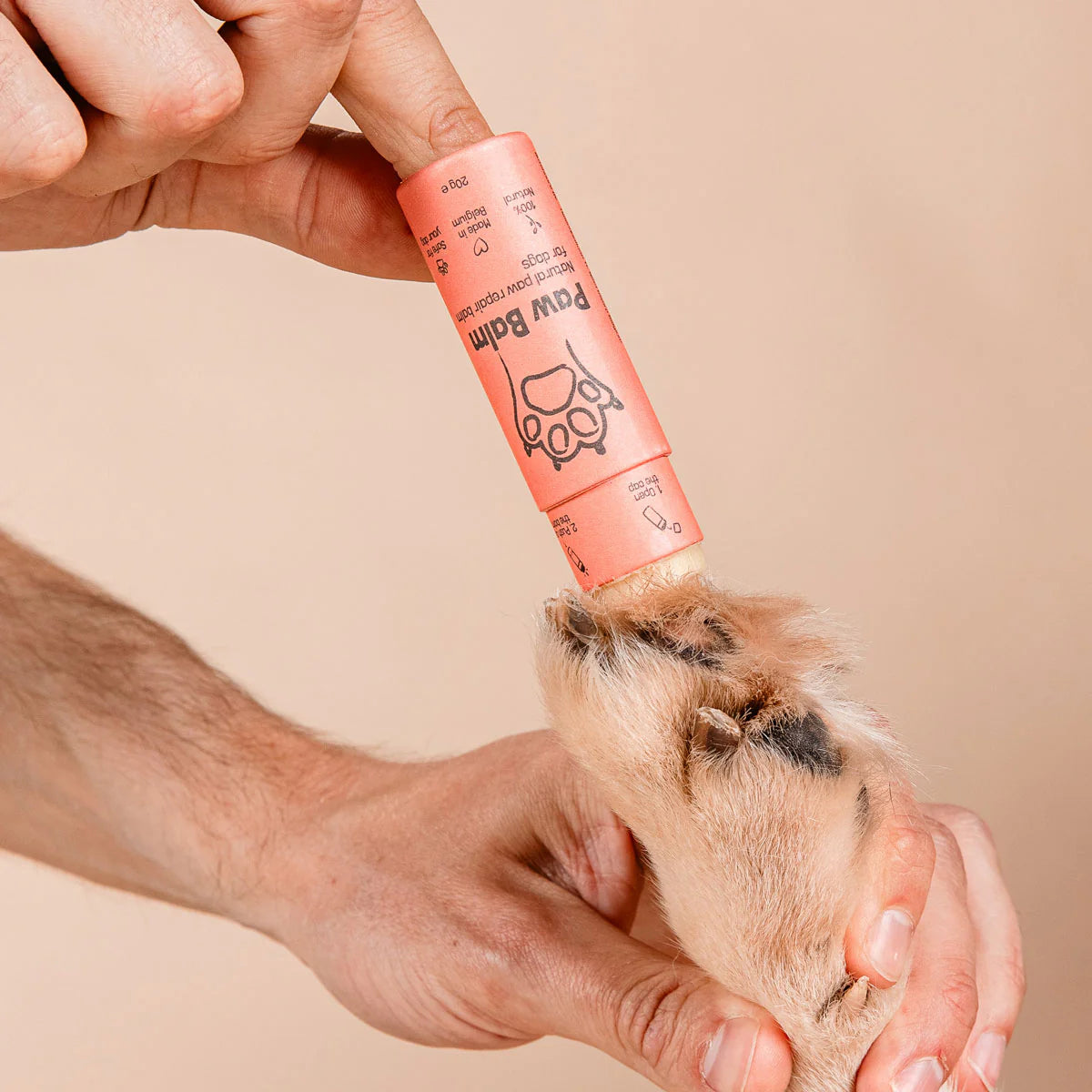 Person applying a pink paw balm to a dog's paw on a beige background