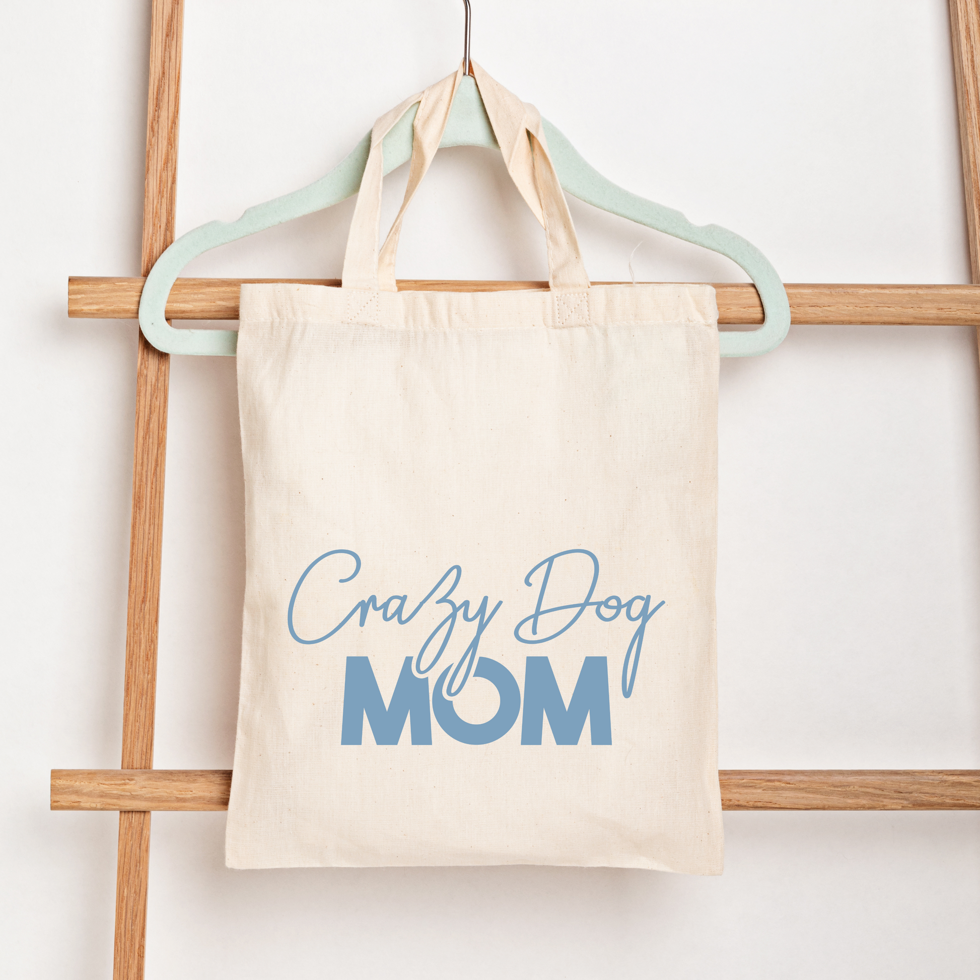 Beige tote bag with 'Crazy Dog Mom' text in blue on a wooden rack.