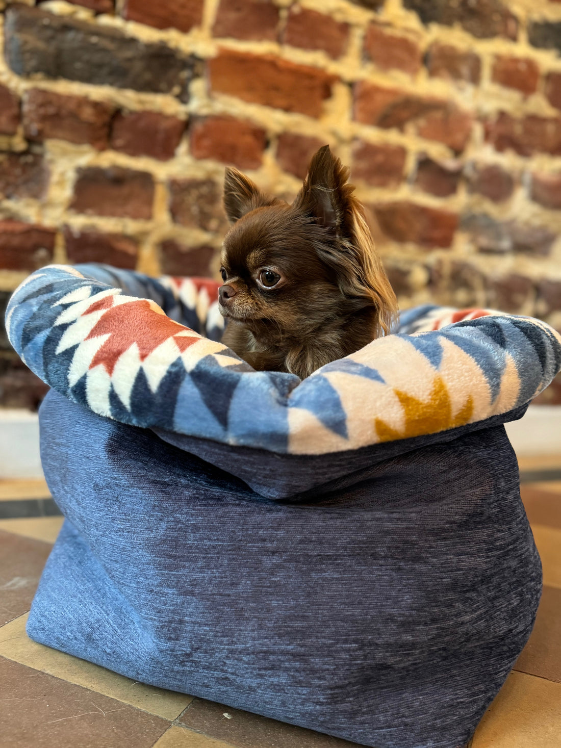 Small dog resting on a colorful geometric-patterned pet bed against a brick wall.