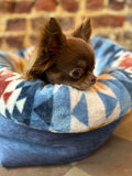 Small brown dog lying on a blue and white patterned pet bed with a brick wall background