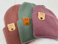 Three knit hats in pink, green, and purple with brand tags on a white background