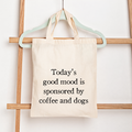 A natural canvas tote bag with black text reading 'Today's good mood is sponsored by coffee and dogs' hanging from a mint green hanger on a wooden ladder against a white wall.