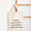 A natural canvas tote bag with black text reading 'Today's good mood is sponsored by coffee and dogs', hanging from a wooden ladder against a white wall.