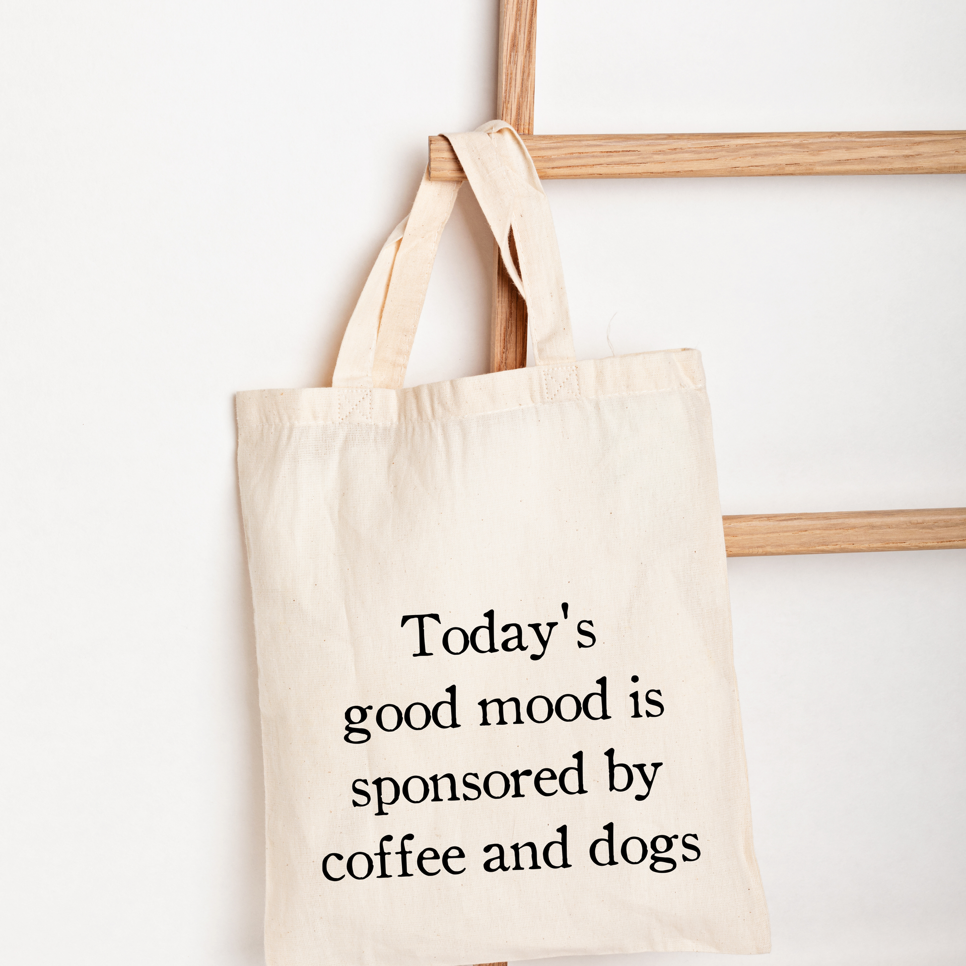 A natural canvas tote bag with black text reading 'Today's good mood is sponsored by coffee and dogs', hanging from a wooden ladder against a white wall.