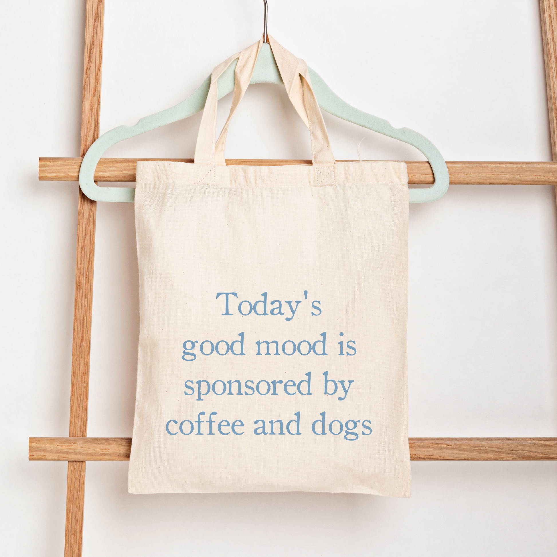 A cream canvas tote bag hanging from a mint green hanger on a wooden ladder, printed with the text 'Today's good mood is sponsored by coffee and dogs' in blue.