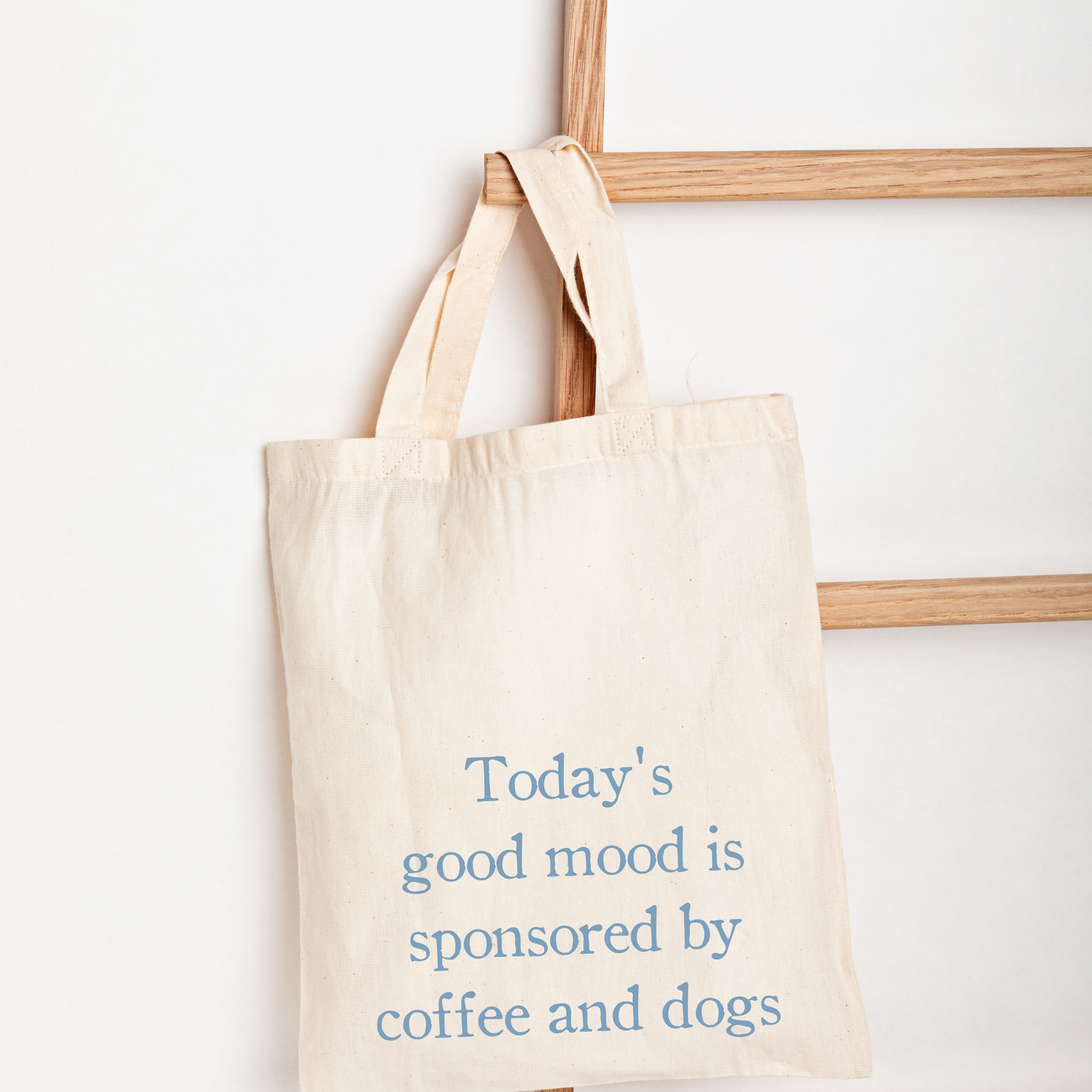 A natural canvas tote bag with blue text reading 'Today's good mood is sponsored by coffee and dogs' hanging from a wooden ladder against a white wall.
