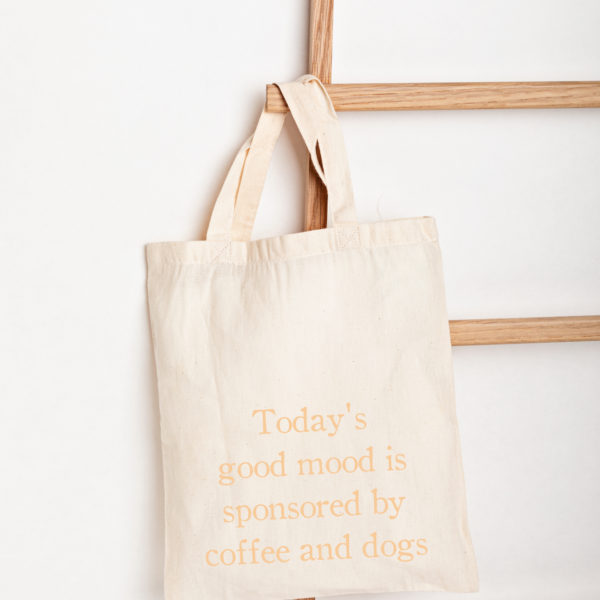 A natural canvas tote bag with orange text reading 'Today's good mood is sponsored by coffee and dogs', hanging from a wooden ladder against a white wall.