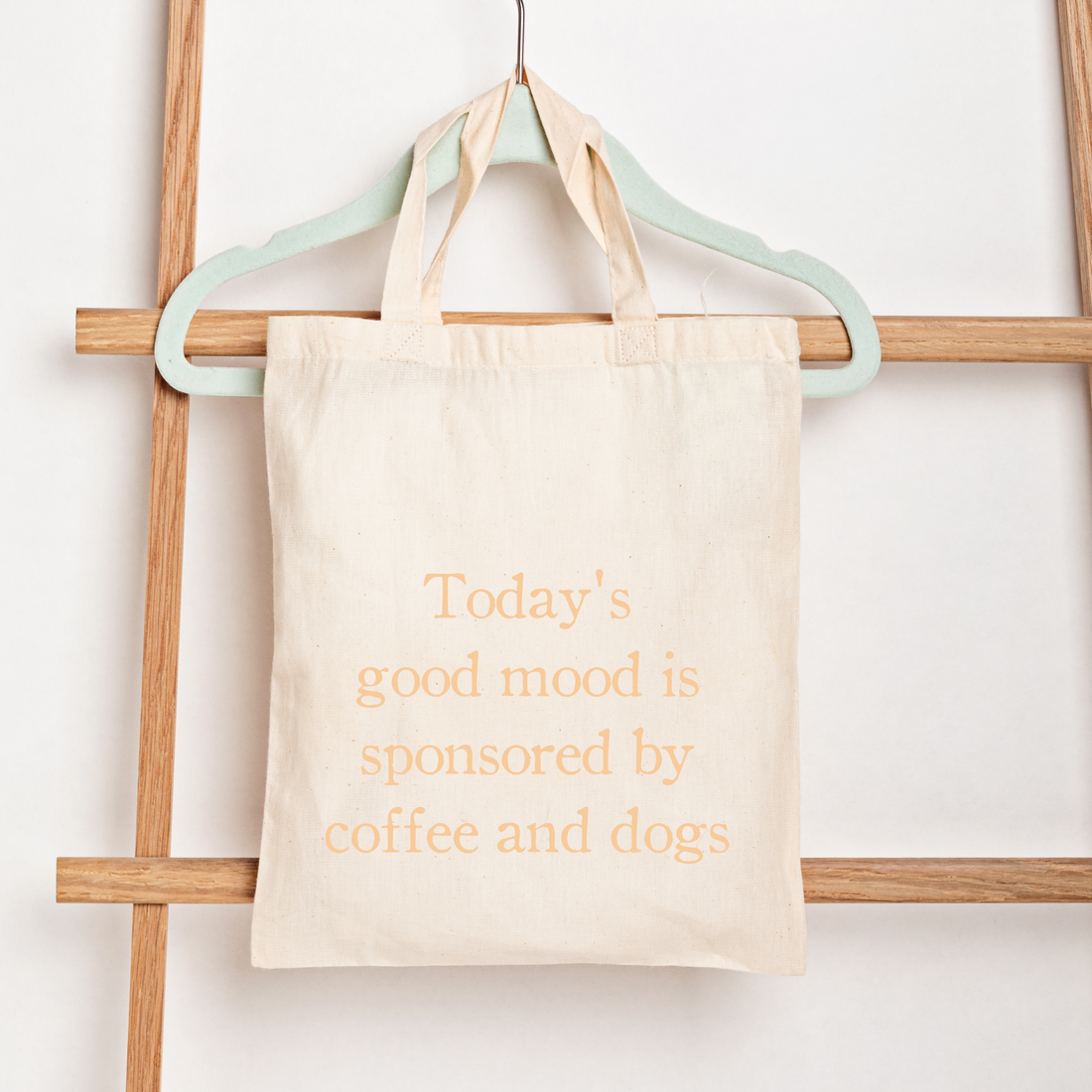 A cream canvas tote bag hanging from a mint green hanger on a wooden ladder against a white wall. The bag features the printed text 'Today's good mood is sponsored by coffee and dogs' in a muted sage green font.