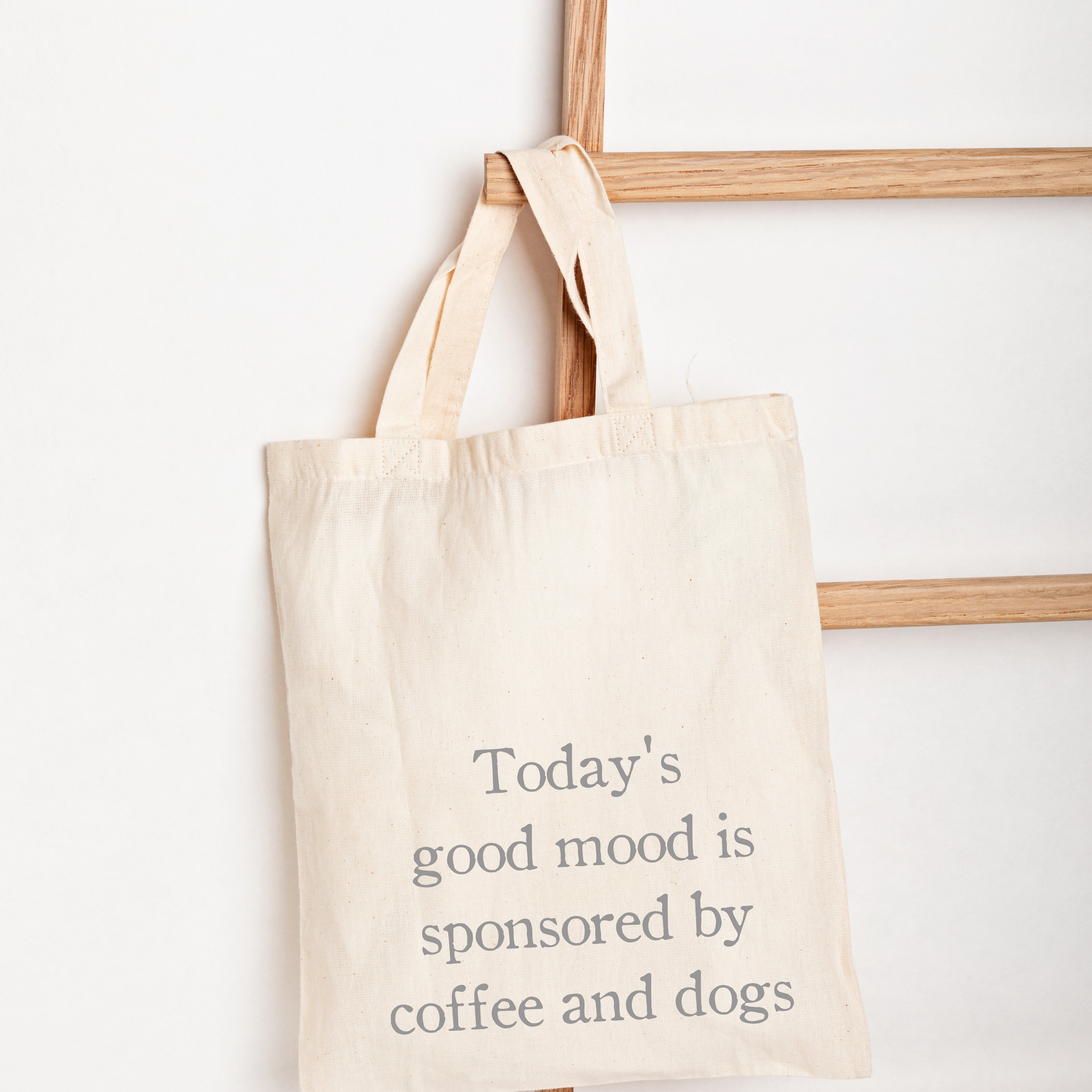A natural canvas tote bag with gray printed text reading 'Today's good mood is sponsored by coffee and dogs', hanging from a wooden ladder against a white wall.
