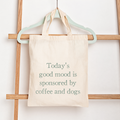 A cream canvas tote bag hanging from a mint green hanger on a wooden ladder against a white wall. The bag features the printed text 'Today's good mood is sponsored by coffee and dogs' in a muted sage green font.