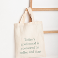 A natural canvas tote bag with the phrase 'Today's good mood is sponsored by coffee and dogs' printed in muted green text, hanging from a wooden ladder against a white wall.