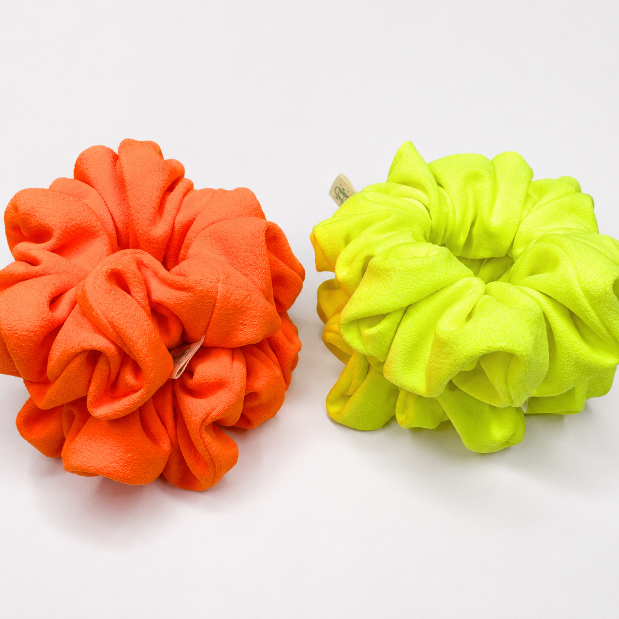 Two fabric hair scrunchies, one in bright orange and the other in neon yellow-green, displayed side-by-side on a clean white background.