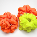 Three fabric scrunchies in bright neon orange and lime green arranged on a clean white background.