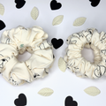 Two cream-colored fabric scrunchies with black cartoon line drawings, displayed on a white background decorated with black and cream paper hearts and leaves.
