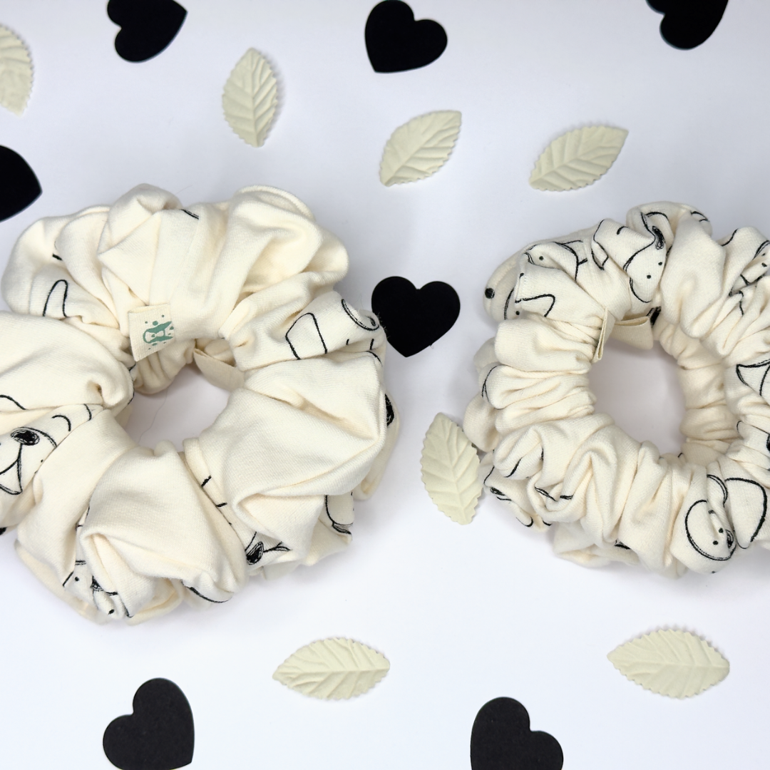 Two cream-colored fabric scrunchies with black cartoon line drawings, displayed on a white background decorated with black and cream paper hearts and leaves.