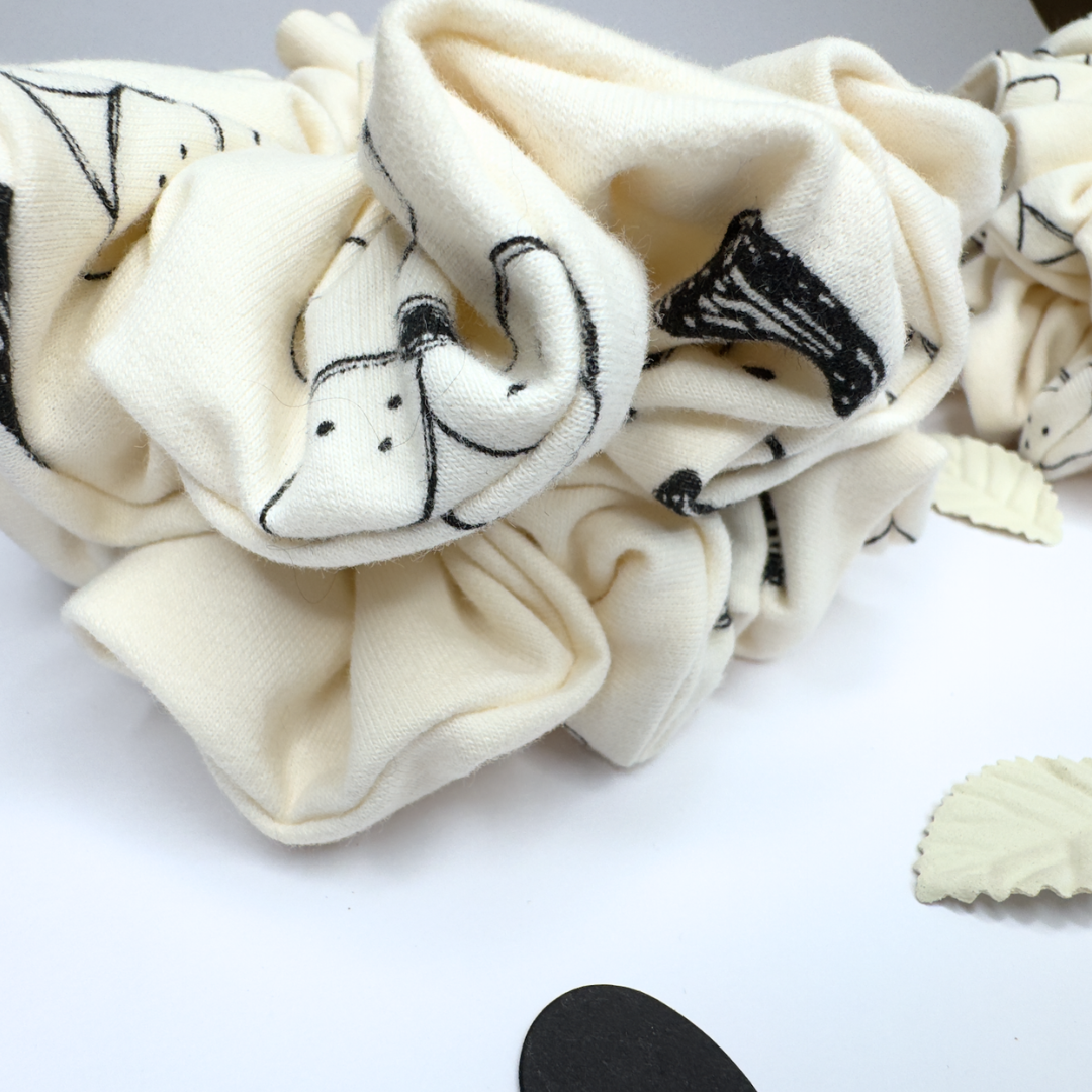 An off-white fleece scrunchie with black line-art cartoon cat illustrations, displayed on a clean white surface.