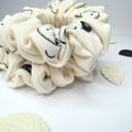 A cream-colored fabric scrunchie with a black cartoon face pattern, displayed on a white surface with decorative leaf-shaped props.