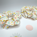 Two fabric hair scrunchies with a white background and pastel multicolor polka dots, displayed on a light gray surface with decorative paper circles and a daisy flower.