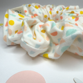 A fabric hair scrunchie with a white base and a colorful confetti dot pattern in pastel yellow, orange, light blue, and mint green, photographed on a white surface.