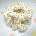 A fabric hair scrunchie with a white base and a colorful polka dot pattern in yellow, orange, and mint green, displayed on a white background with subtle decorative paper cutouts.