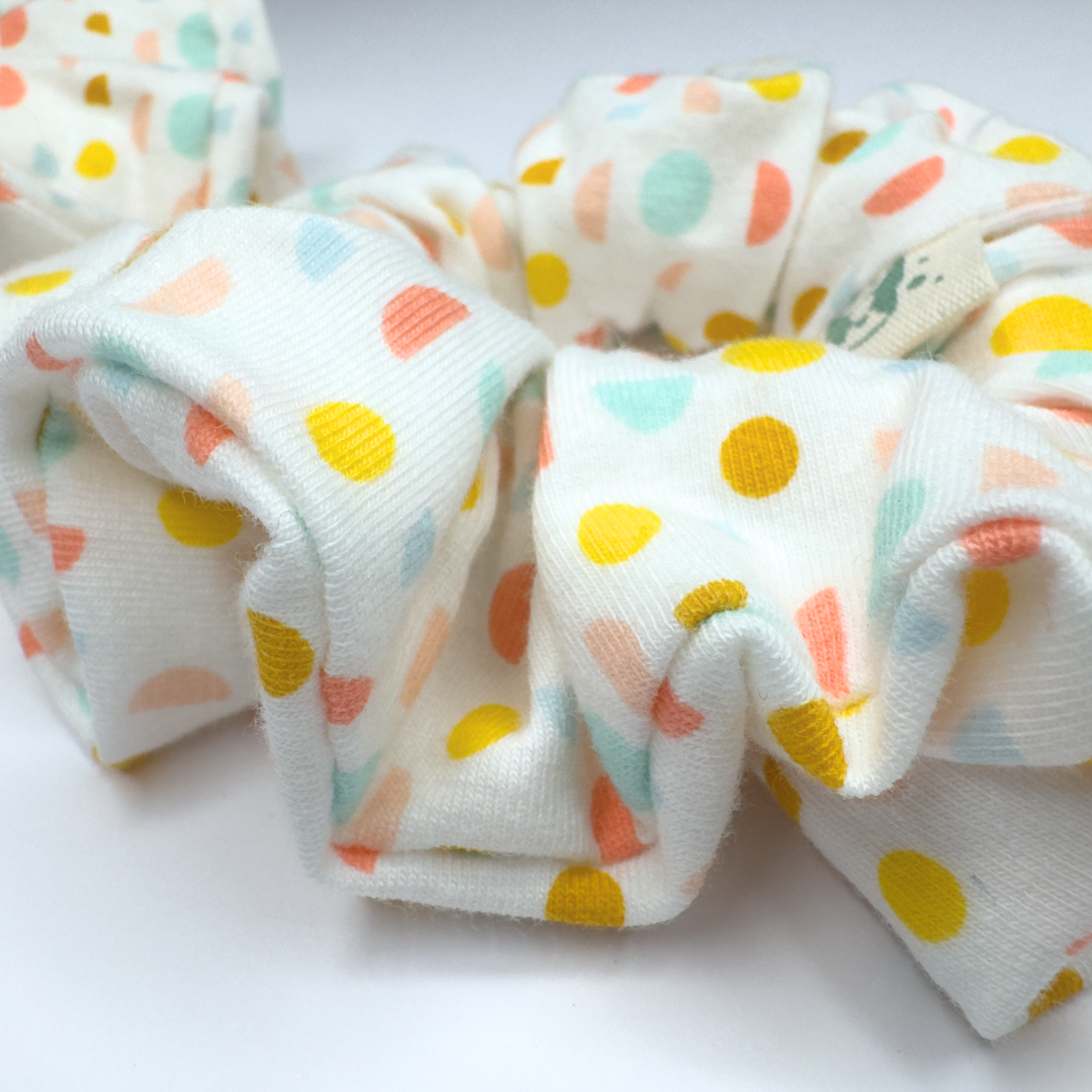 A fabric hair scrunchie with a white base and a colorful polka dot pattern in yellow, orange, and light blue, shown in a close-up shot on a white background.