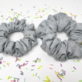 Two gray fabric scrunchies with a textured linen-like weave, displayed on a white background with scattered pastel sprinkle decorations.