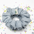 A light blue-gray fabric scrunchie with a small beige brand tag featuring a green bear logo, set against a white background with scattered colorful sprinkles.