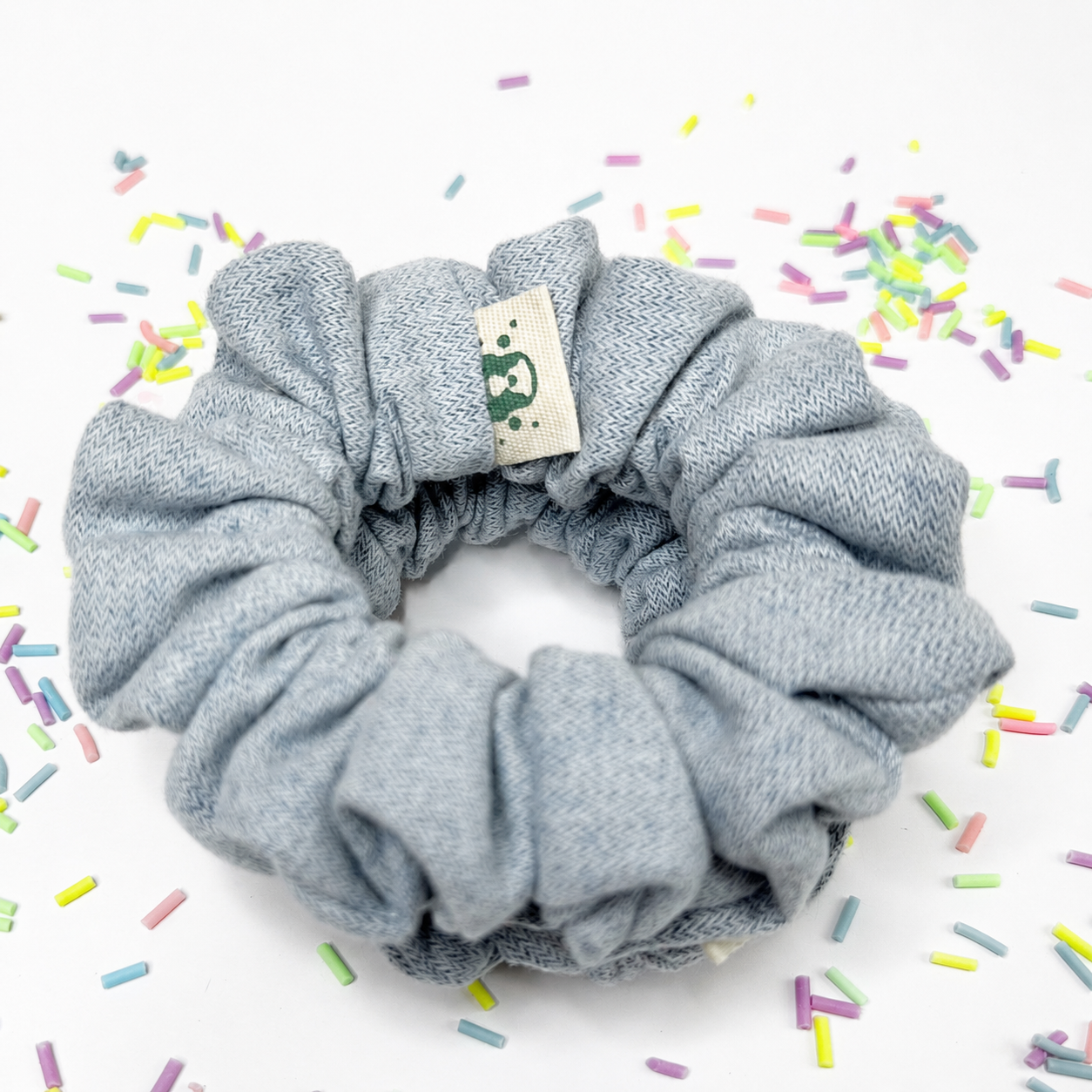 A light blue-gray fabric scrunchie with a small beige brand tag featuring a green bear logo, set against a white background with scattered colorful sprinkles.