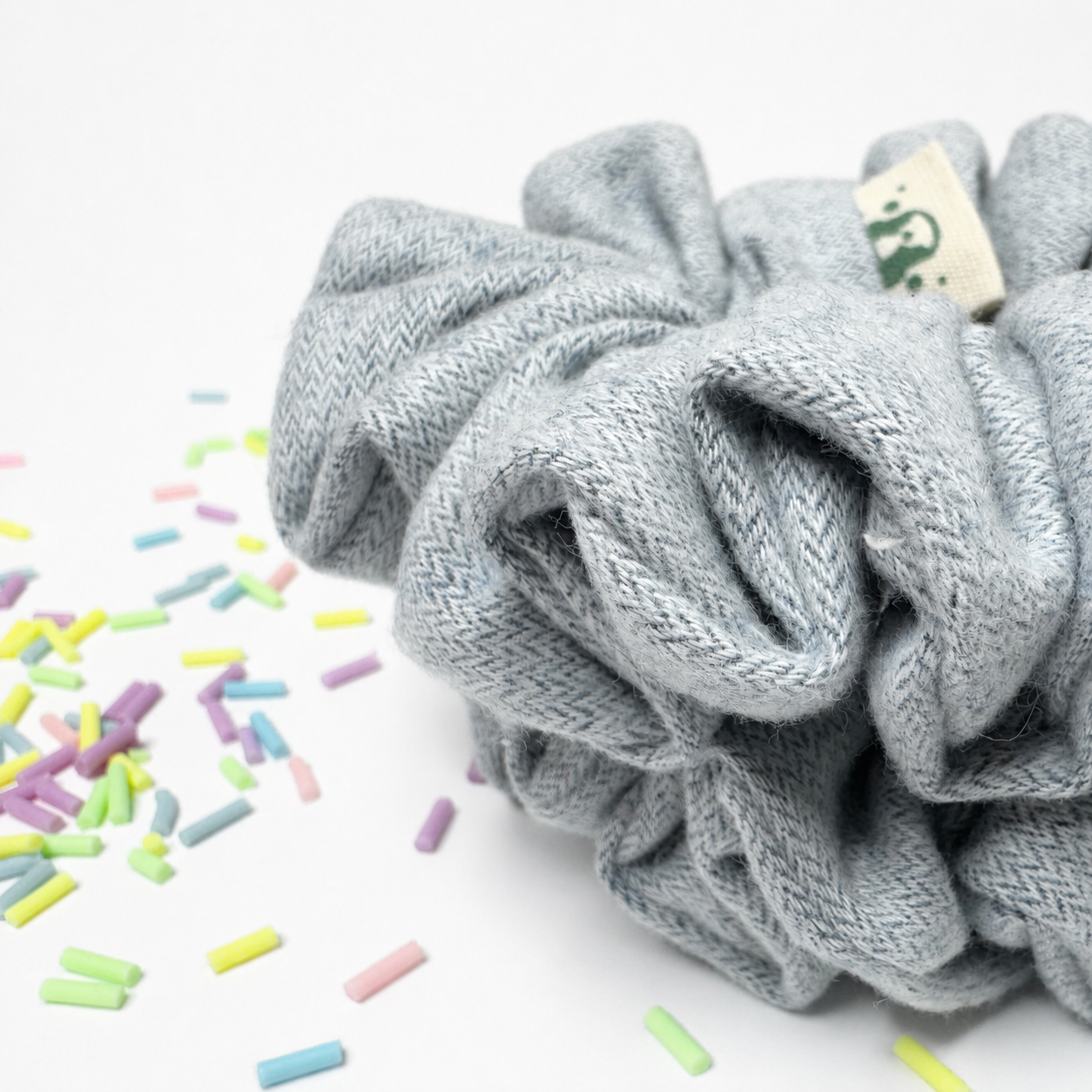 A light gray herringbone knit scrunchie with a small cream fabric tag, photographed on a white surface with scattered pastel sprinkles in the foreground.