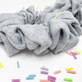 A light gray textured fabric scrunchie with a woven herringbone pattern, displayed on a white surface with colorful pastel chalk sticks scattered around.