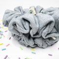 A light gray herringbone-patterned fabric scrunchie with a small cream paper tag, displayed on a white surface with colorful sprinkle props.