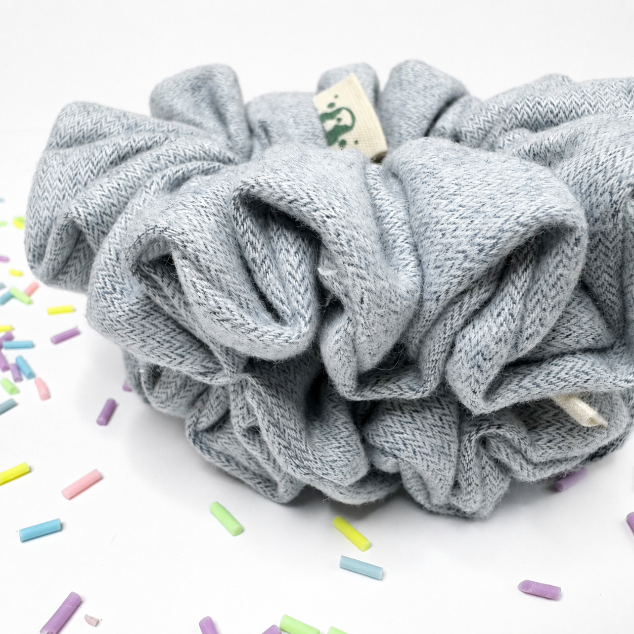 A light gray herringbone-patterned fabric scrunchie with a small cream paper tag, displayed on a white surface with colorful sprinkle props.