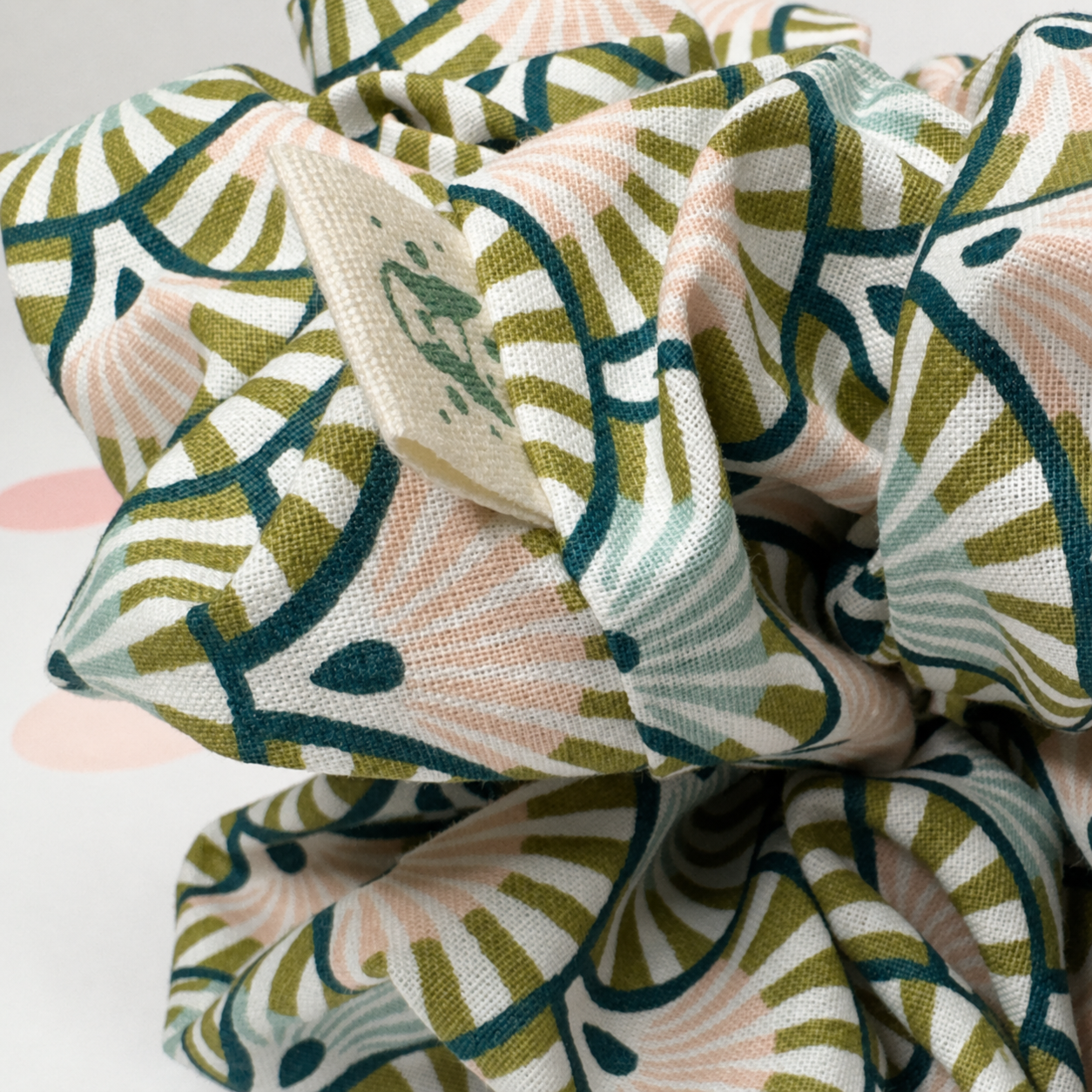 A close-up of a fabric scrunchie with a botanical leaf pattern in sage green, teal, and soft peach on a white background.