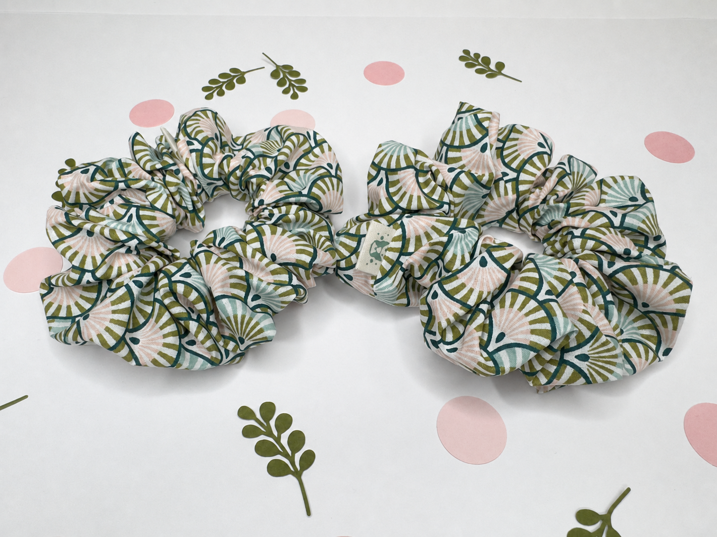 Two fabric hair scrunchies with a green and beige fan pattern, displayed on a white background with pink polka dots and green leaf accents.