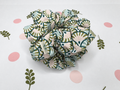 A fabric hair scrunchie with a green, beige, and white geometric fan pattern, displayed on a white background decorated with pink polka dots and green leaf illustrations.