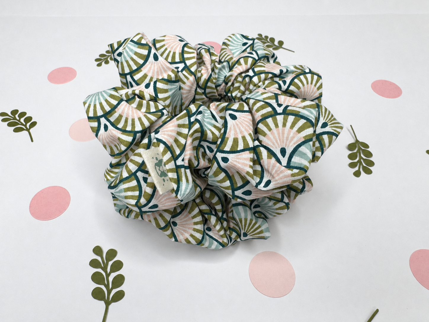 A fabric hair scrunchie with a green, beige, and white geometric fan pattern, displayed on a white background decorated with pink polka dots and green leaf illustrations.