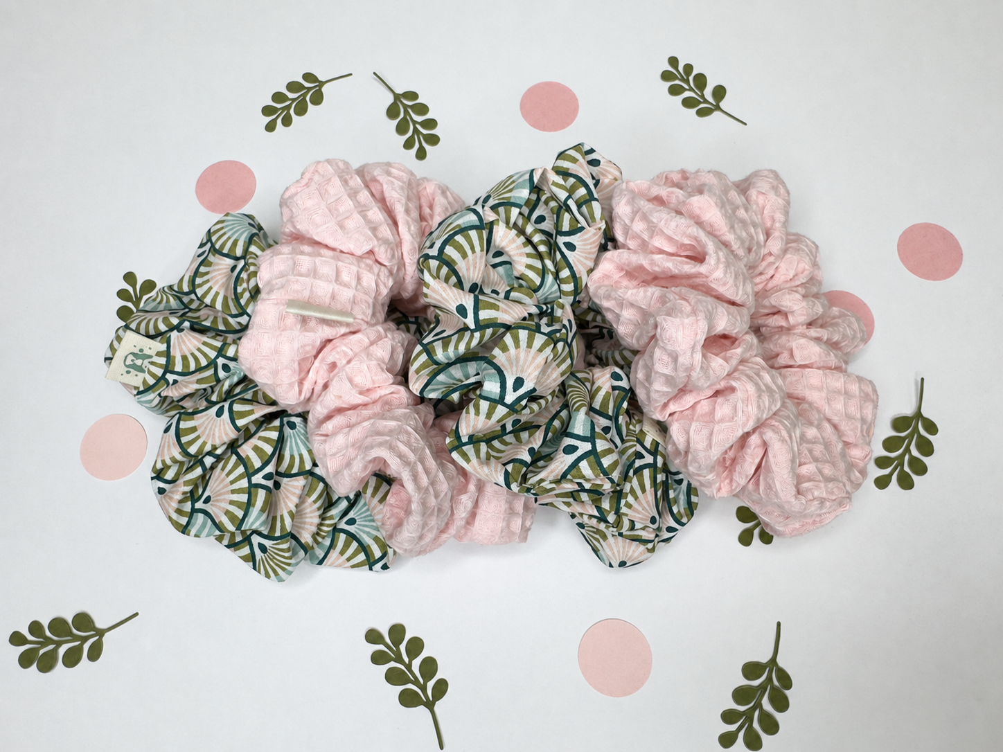 Three fabric hair scrunchies arranged on a white background with decorative pink polka dots and green leaf prints. Two scrunchies are light pink, and one has a green and white palm leaf pattern.