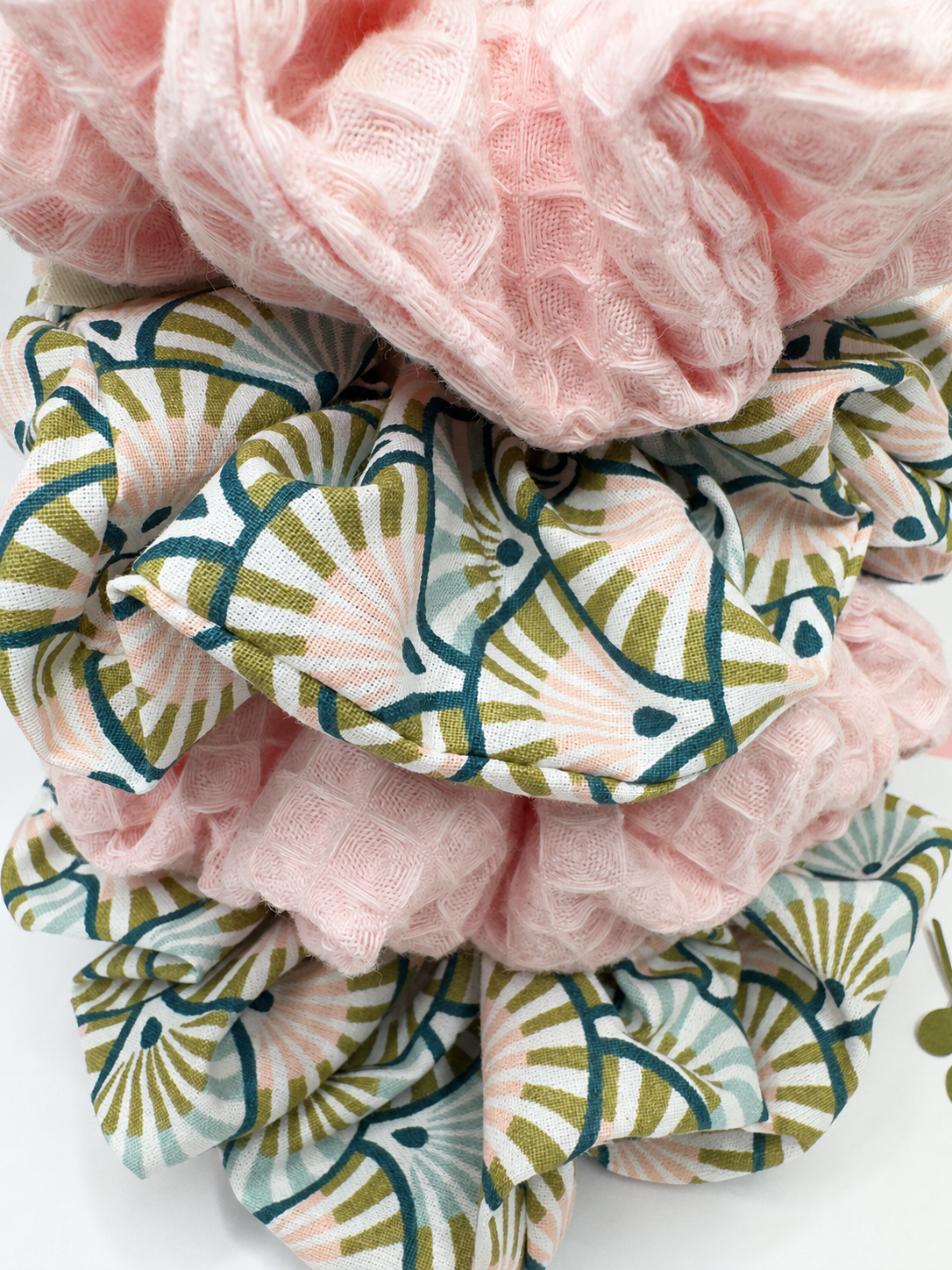 A stack of fabric hair scrunchies, including one solid light pink gauze and two patterned scrunchies with a green and white fan-like geometric design.