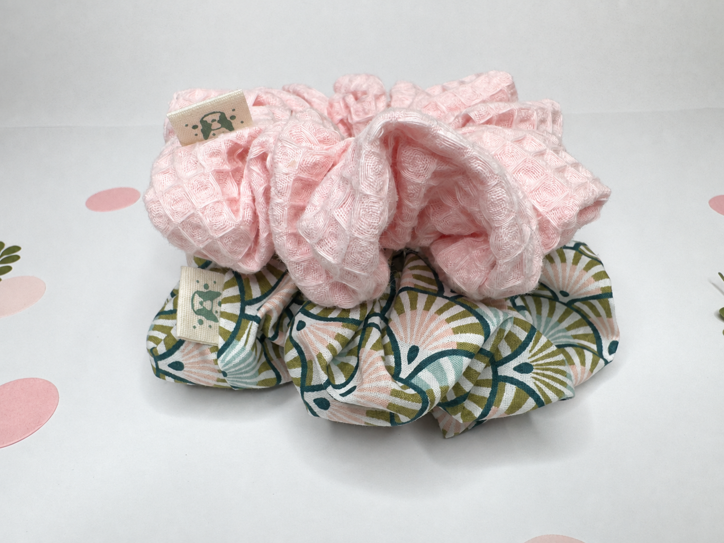 Two fabric hair scrunchies stacked on a white surface. The top scrunchie is light pink with a textured waffle pattern, and the bottom scrunchie features a green, beige, and white scallop pattern.
