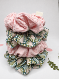 A stack of three textured fabric hair scrunchies on a white background. The top scrunchie is solid light pink, while the bottom two feature a green, beige, and white fan-like geometric pattern.